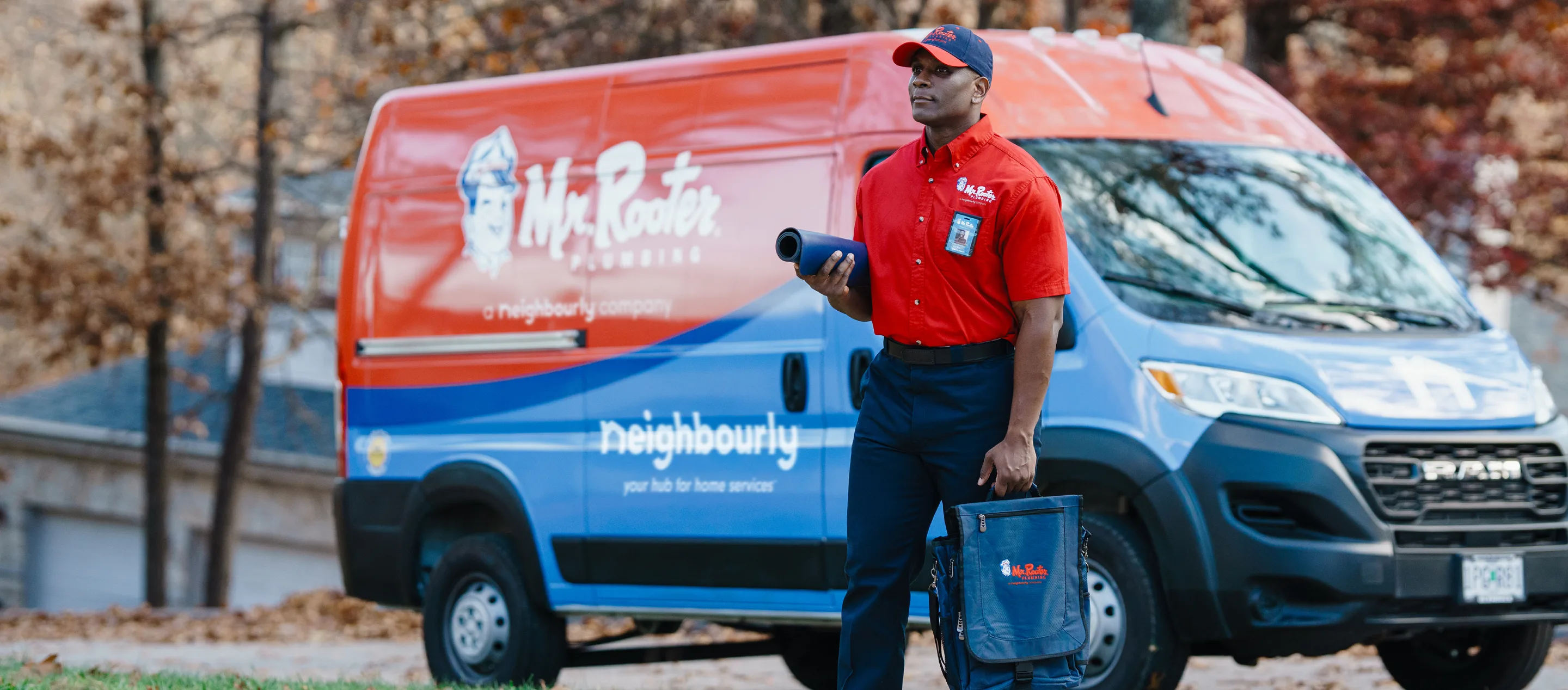 Mr. Rooter technician carrying mat and utility bag in front of branded red work van.