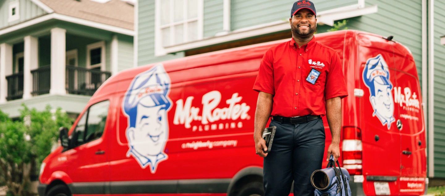 Mr. Rooter technician carrying mat and utility bag in front of branded red work van.