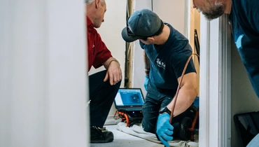 A plumber inspecting a plumbing system while referencing a laptop during a residential repair.