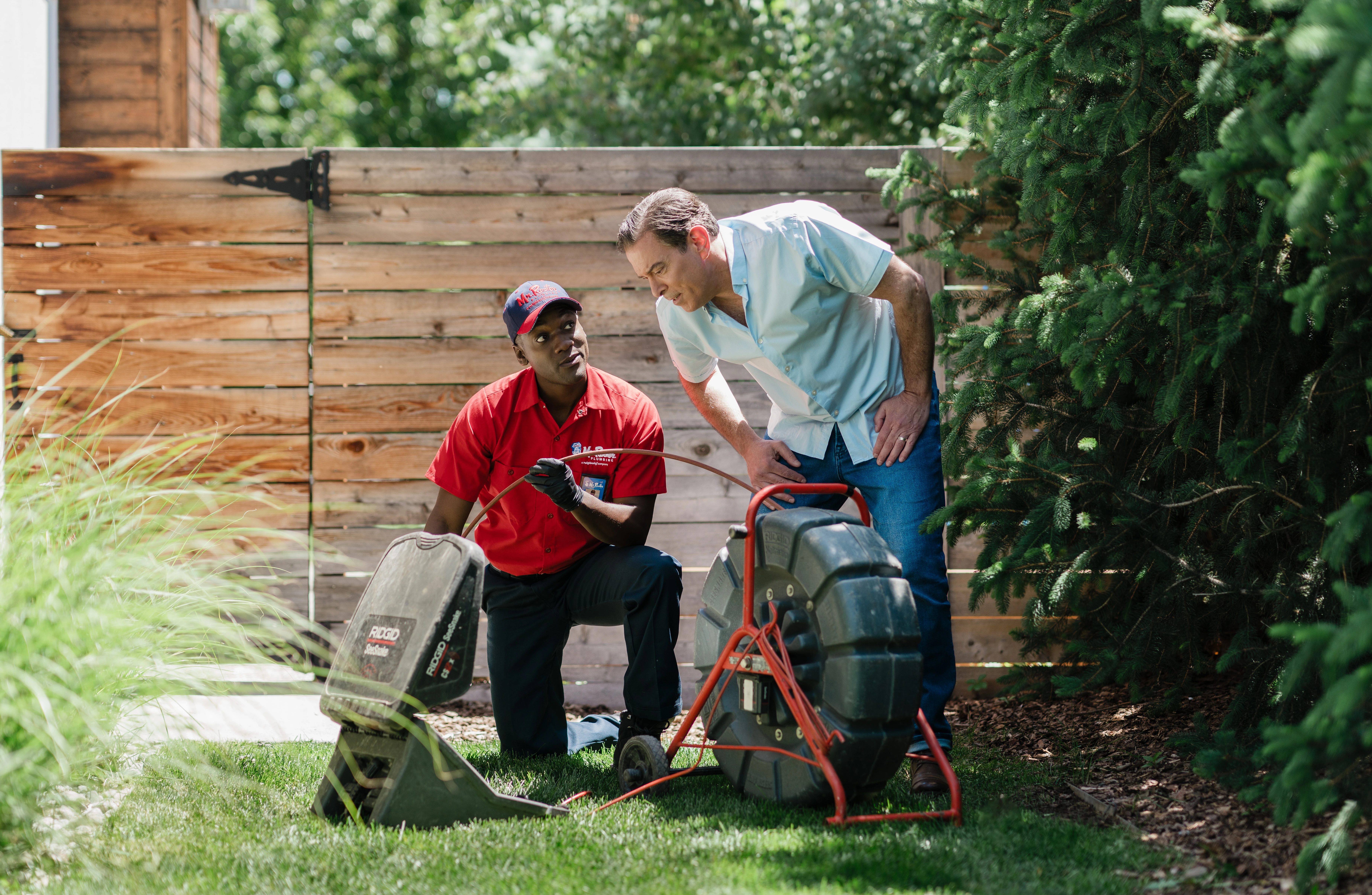 Mr. Rooter Plumbing service professional demonstrating a sewer line camera inspection to a homeowner outdoors, showing the diagnostic equipment used to identify sewer line issues.