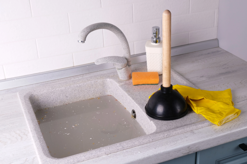 Plunger and yellow rubber gloves next to a clogged sink drain, representing simple DIY plumbing jobs for Canadian homeowners.