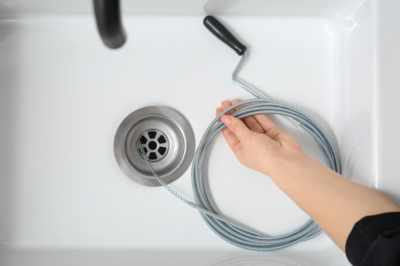 Hand using a plumber’s drain snake near a clogged bathroom sink during simple DIY plumbing repair in Canada.