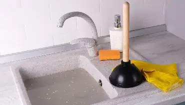 Plunger and yellow rubber gloves next to a clogged sink drain, representing simple DIY plumbing jobs for Canadian homeowners.